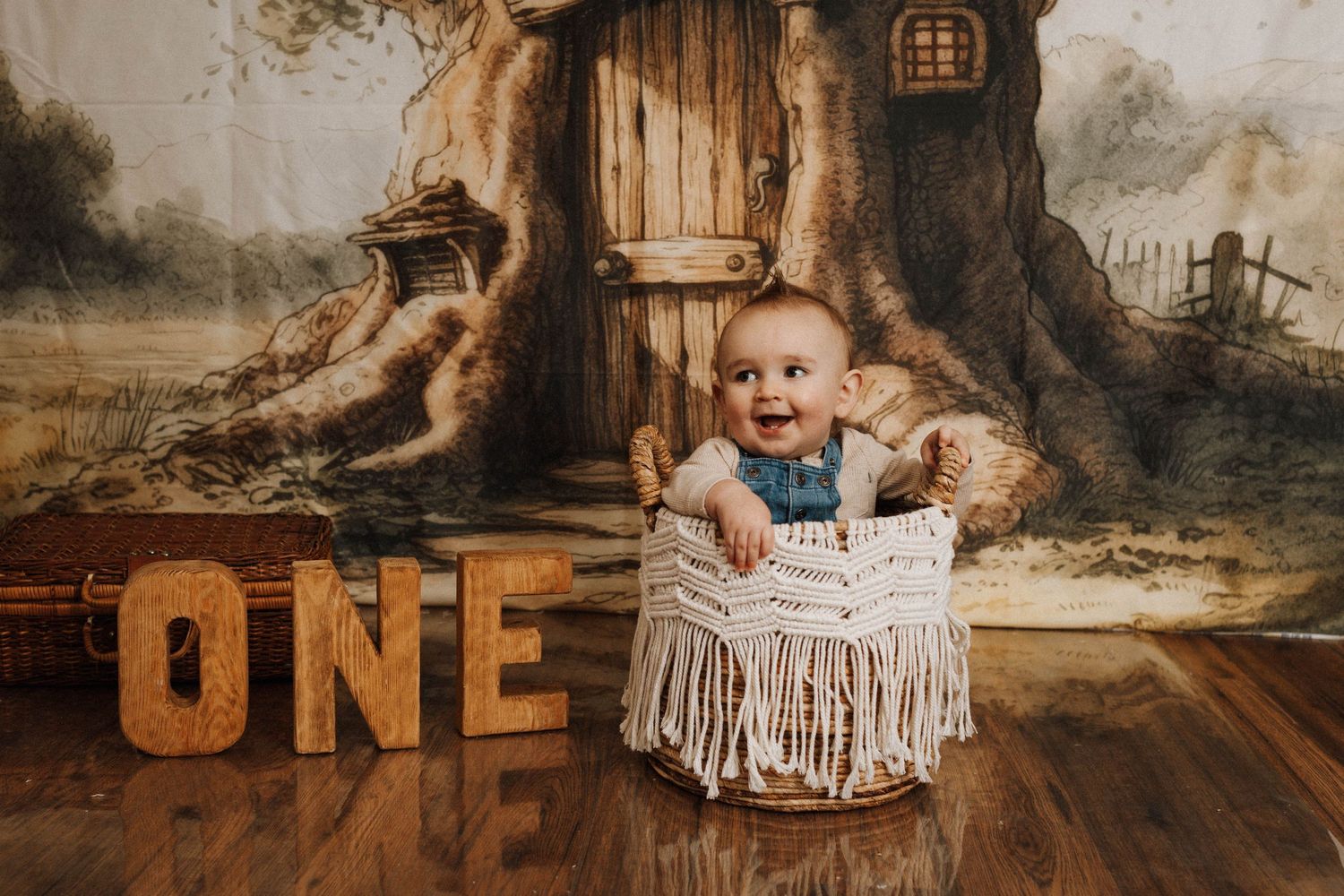 Baby in denim overalls sitting in macrame basket against enchanted forest backdrop.