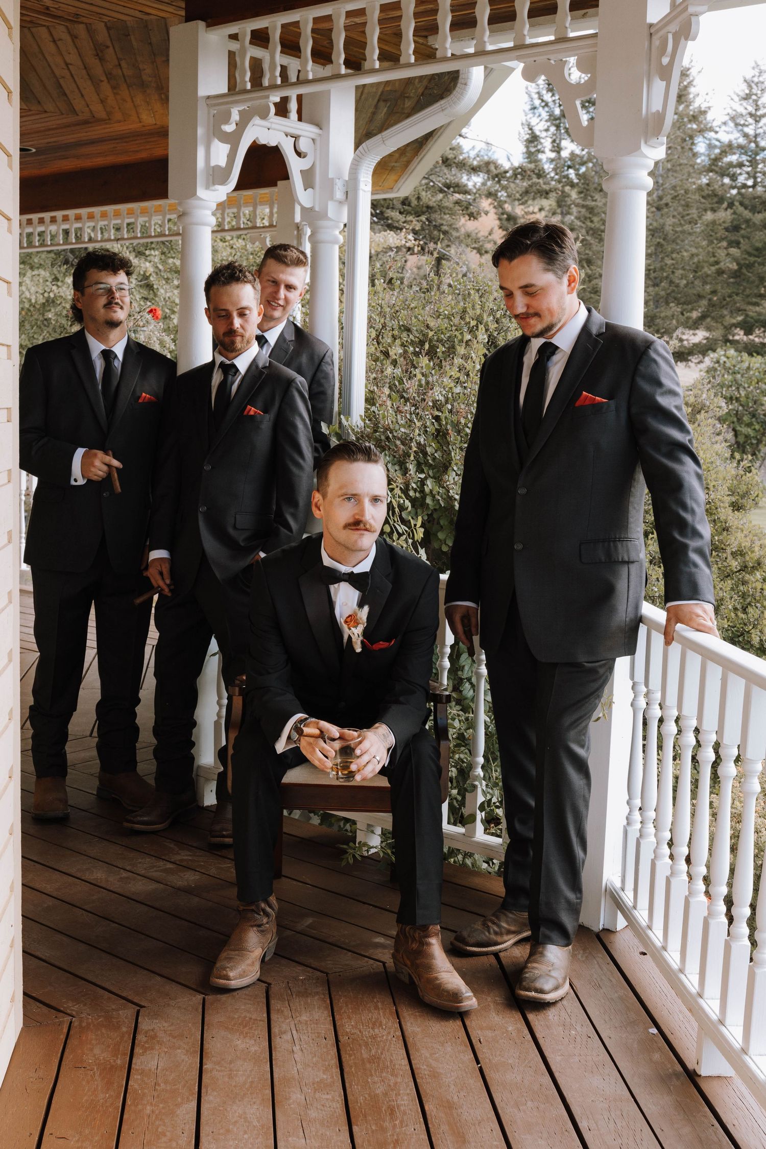 Groomsmen in black suits with red accents gather on a white porch railing during a wedding celebration.