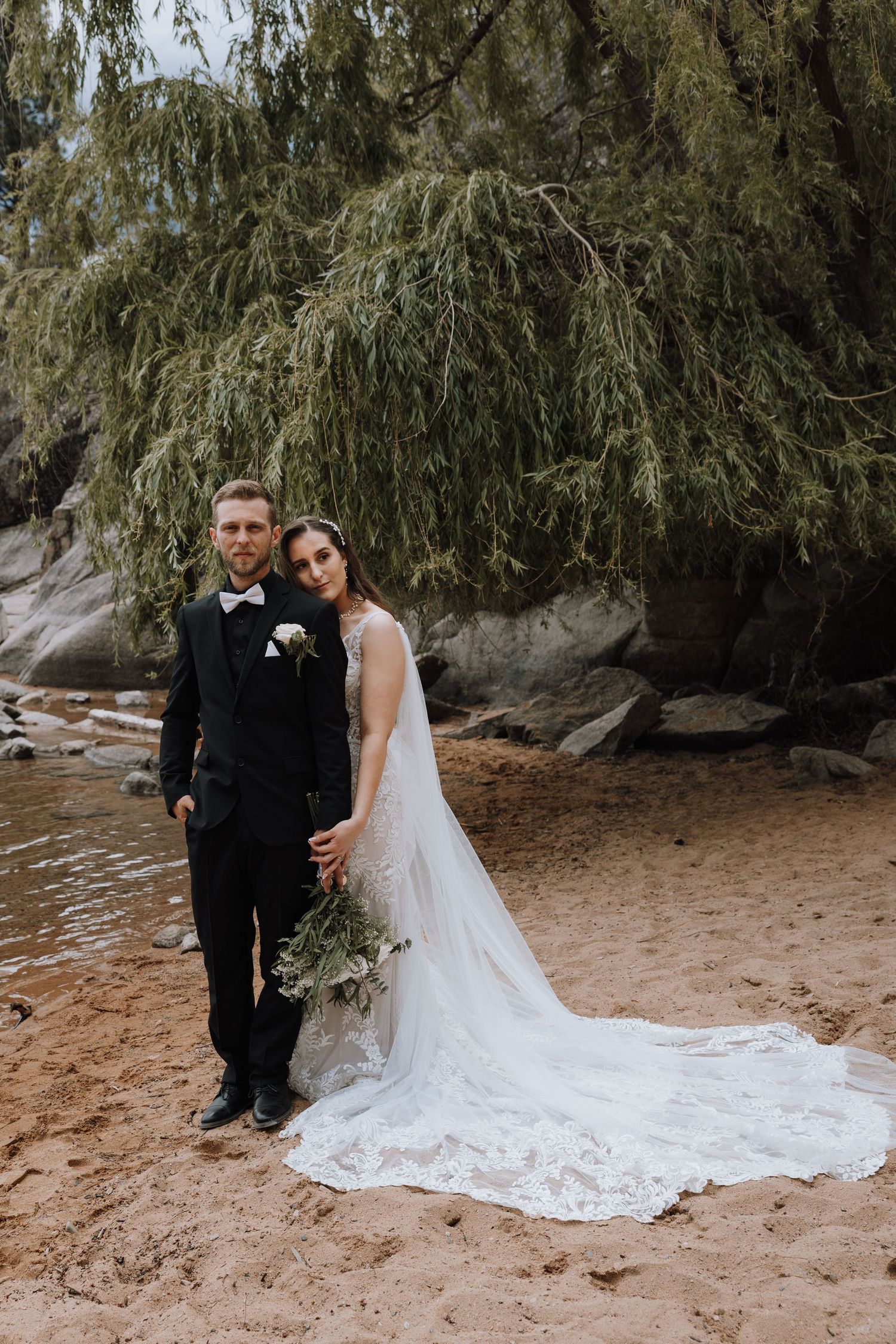Bride and groom posing on sandy beach under weeping willow tree during outdoor wedding photoshoot.