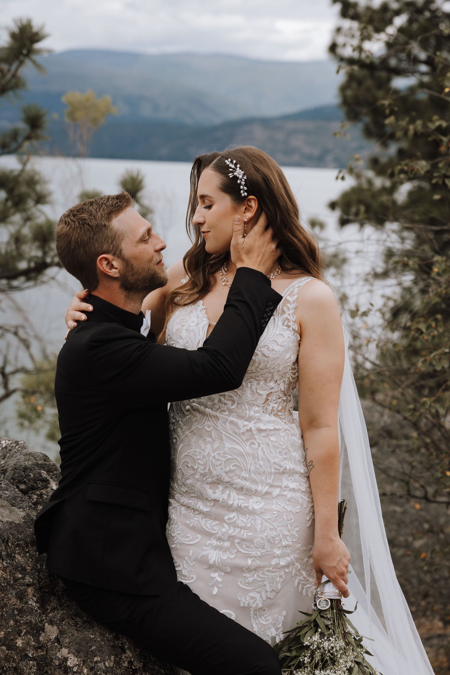 A groom in a black suit tenderly holds his bride's face as she holds a bouquet, with a scenic lake and mountains behind them.