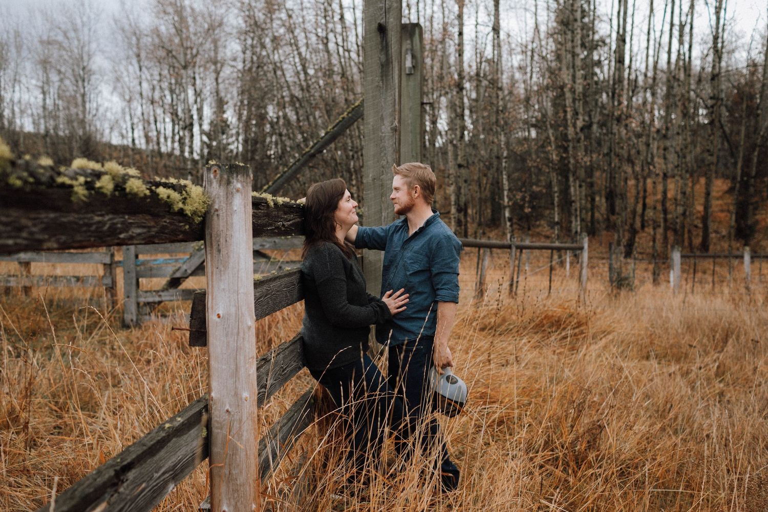 A romantic couple shares a tender moment beside a rustic wooden fence in a golden autumn field.