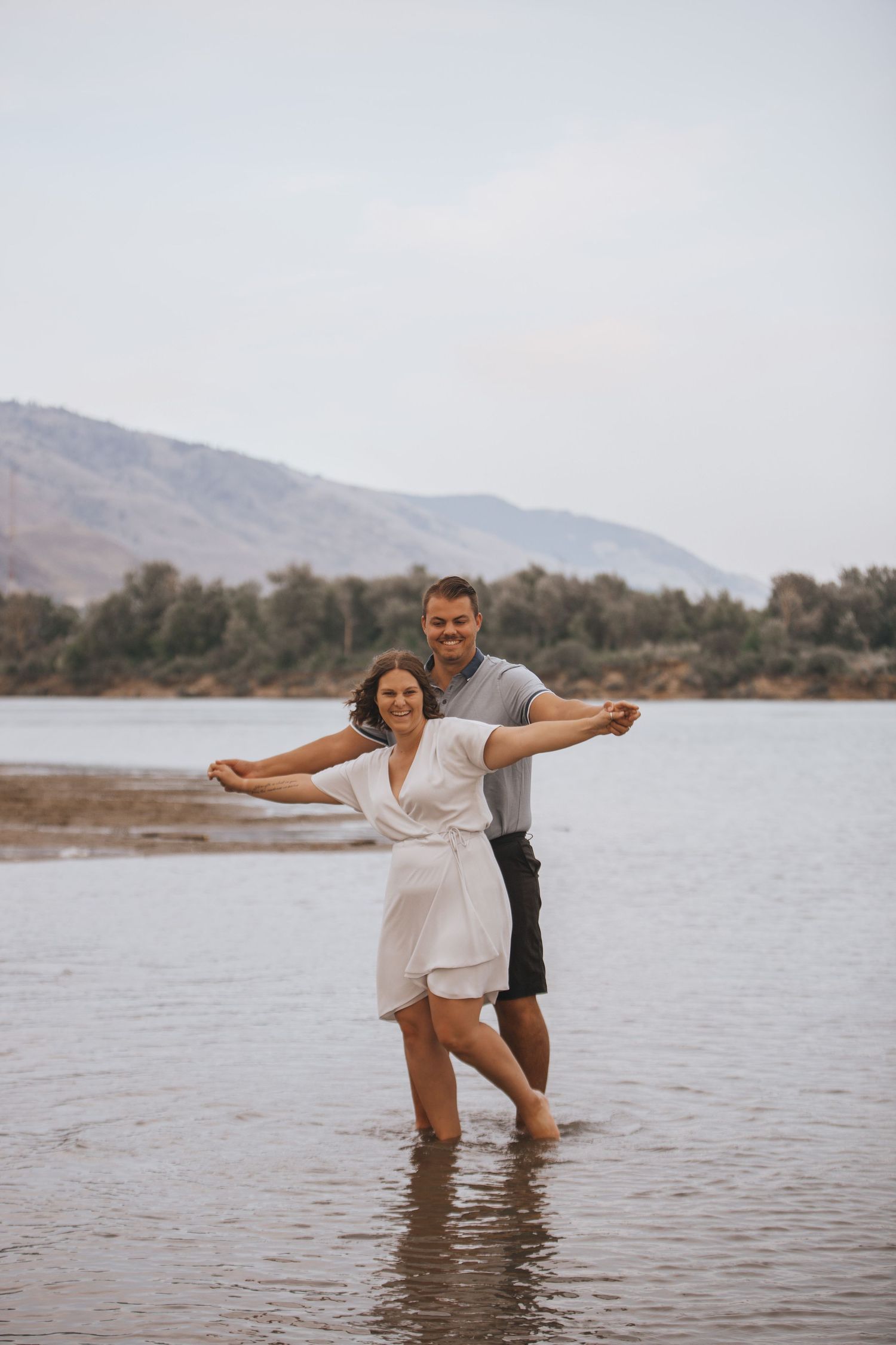 A couple spreads their arms while dancing in shallow water near a mountainous shoreline.