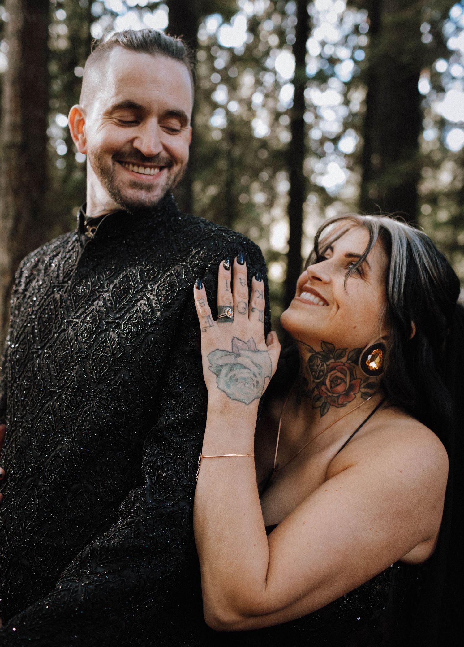 Engaged couple showing off ring in a forest setting with visible tattoos and joyful expressions.