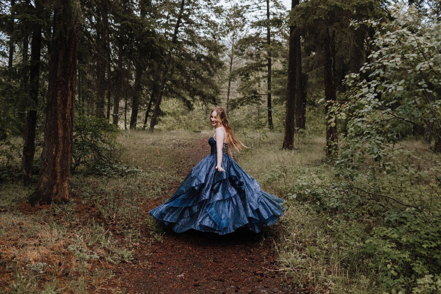 A sparkling navy blue ballgown stands out against a woodland forest path backdrop.