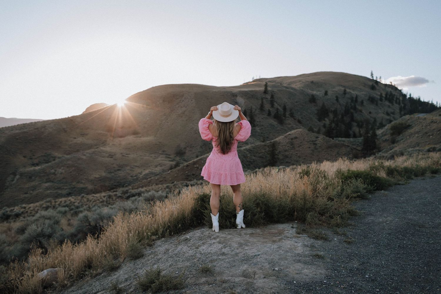 A girl in a pink dress and hat stands on a mountain trail at sunset watching the sun peek over distant hills.