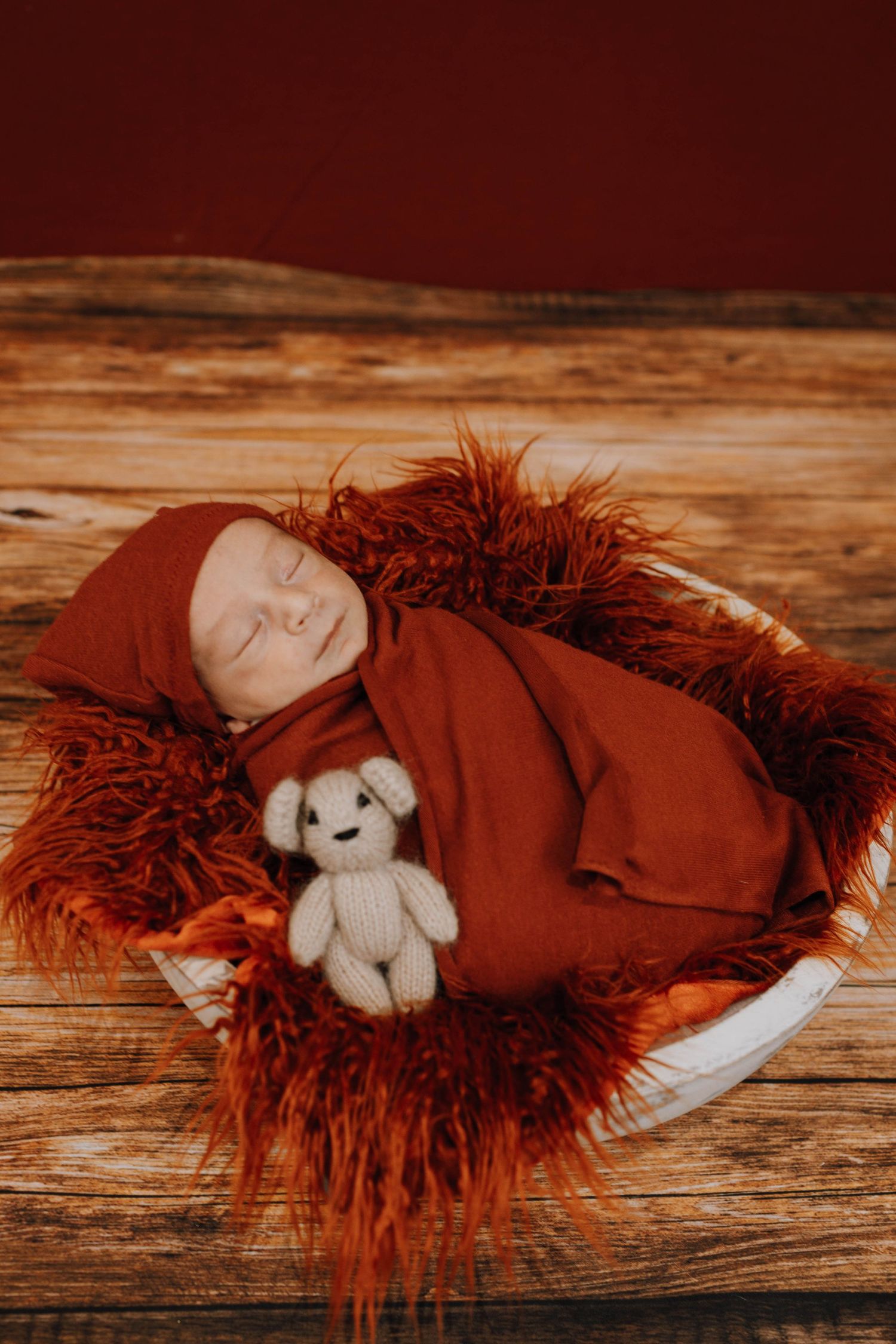 Baby sleeping on red fur blanket with small white teddy bear stuffed animal companion.