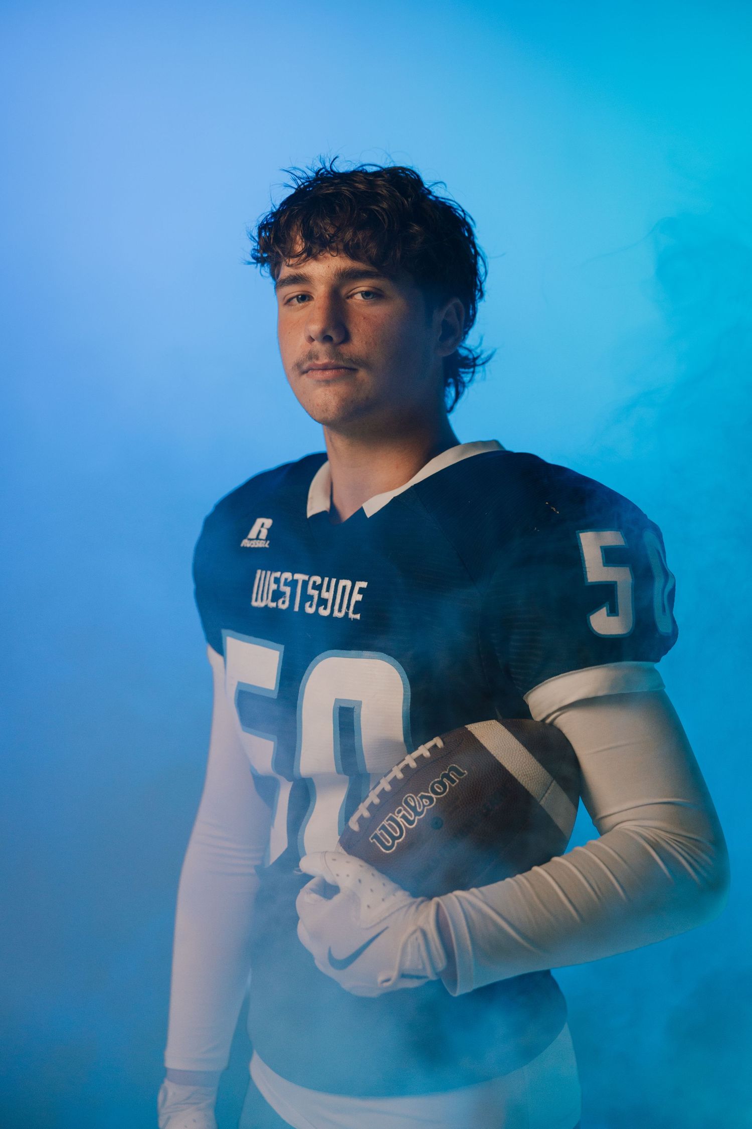 Young football player in Westsyde #50 jersey holds Wilson ball against blue smoky background.