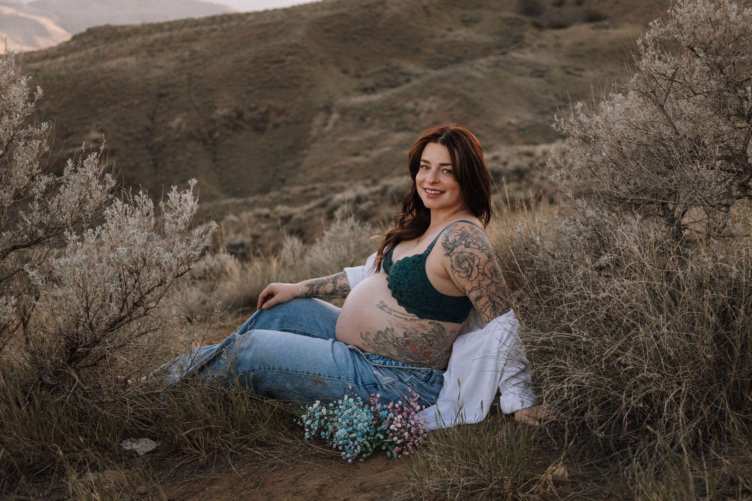 A pregnant woman in blue jeans sits on a hillside surrounded by sage brush in warm golden light.