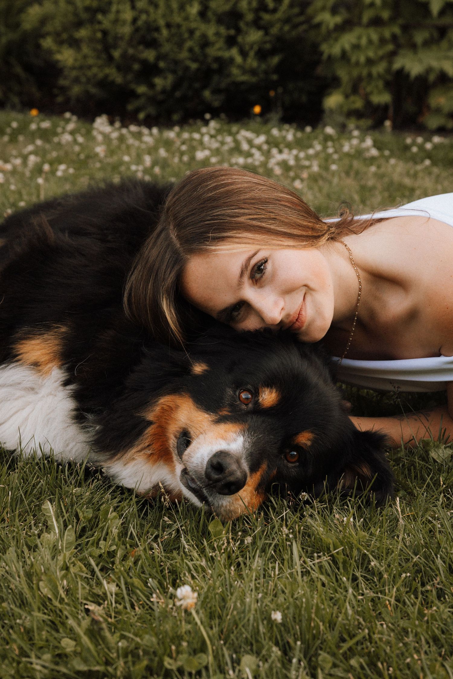 A young woman in white smiles while lying on grass cuddling her tri-color Bernese Mountain Dog outdoors.