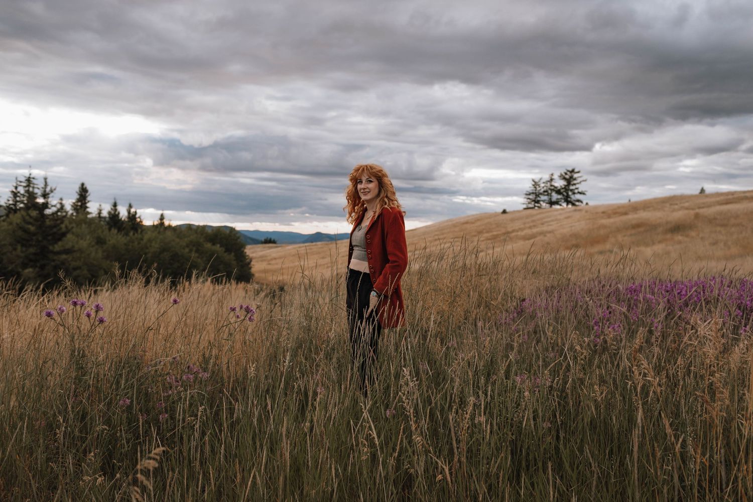 A figure in a rust-colored coat stands in a golden meadow under dramatic cloudy skies.