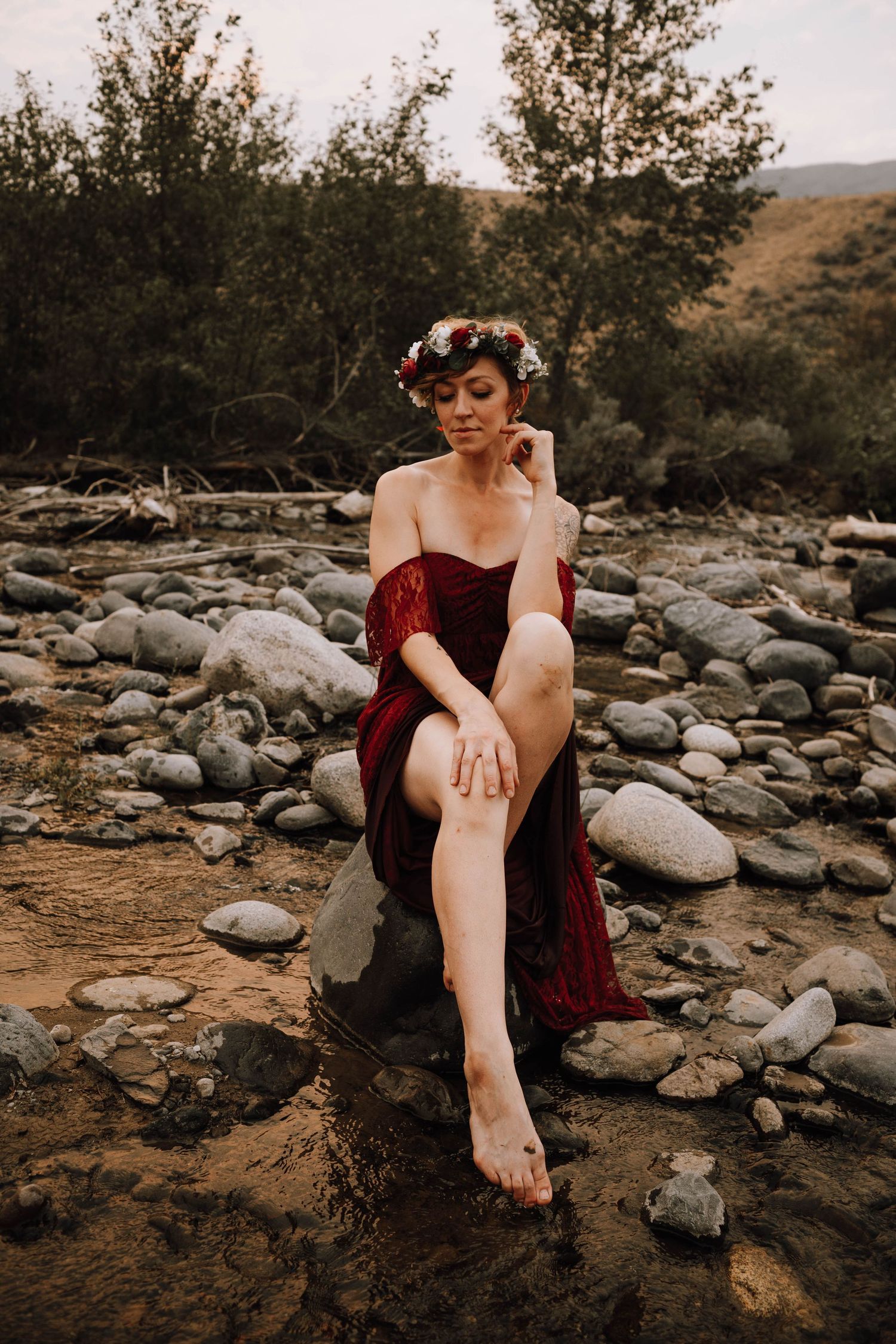 Vintage-inspired portrait with floral crown and burgundy dress against rocky natural backdrop.