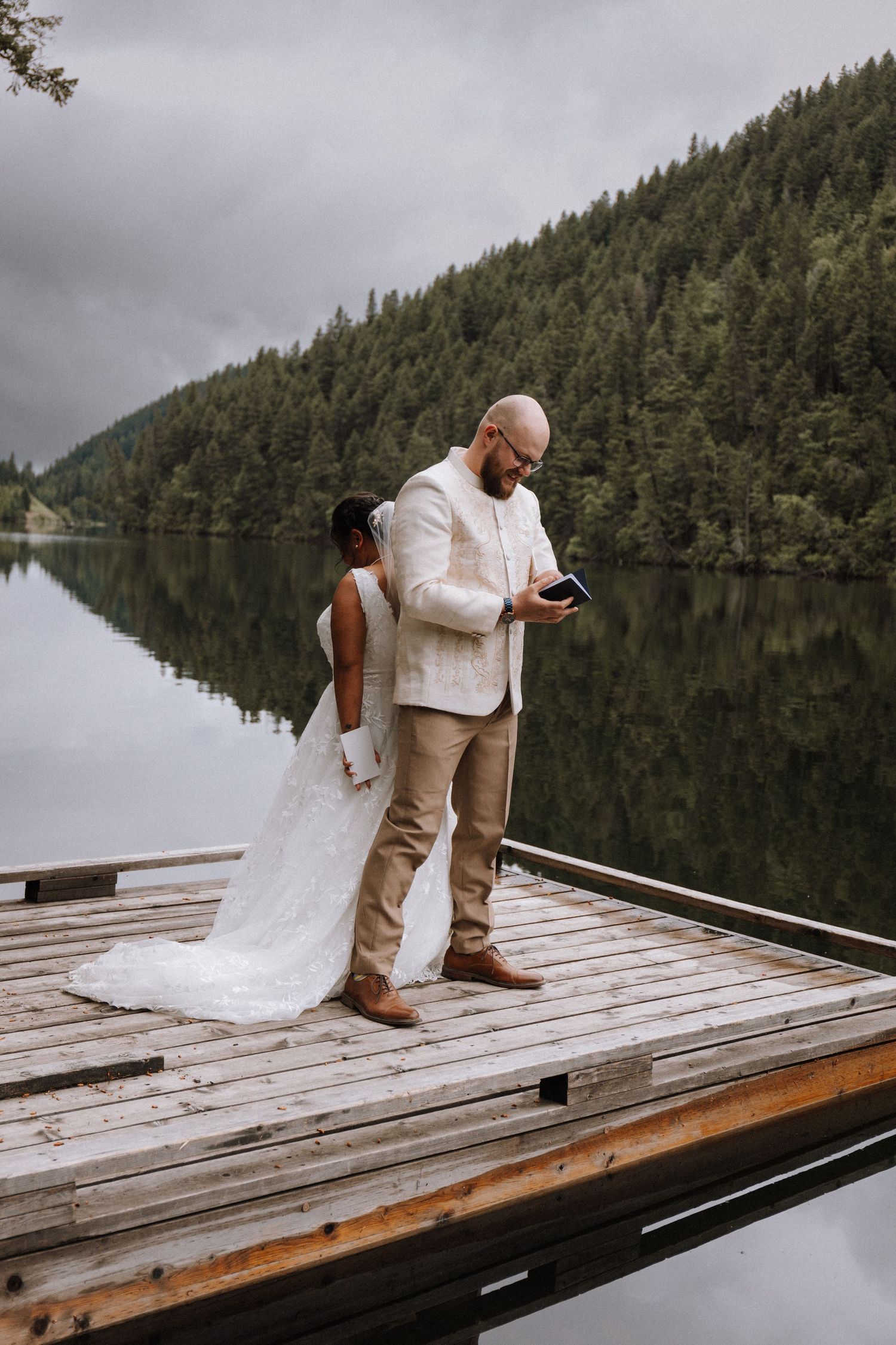 A couple kneels on a wooden dock surrounded by evergreen trees and a serene lake in the Pacific Northwest.