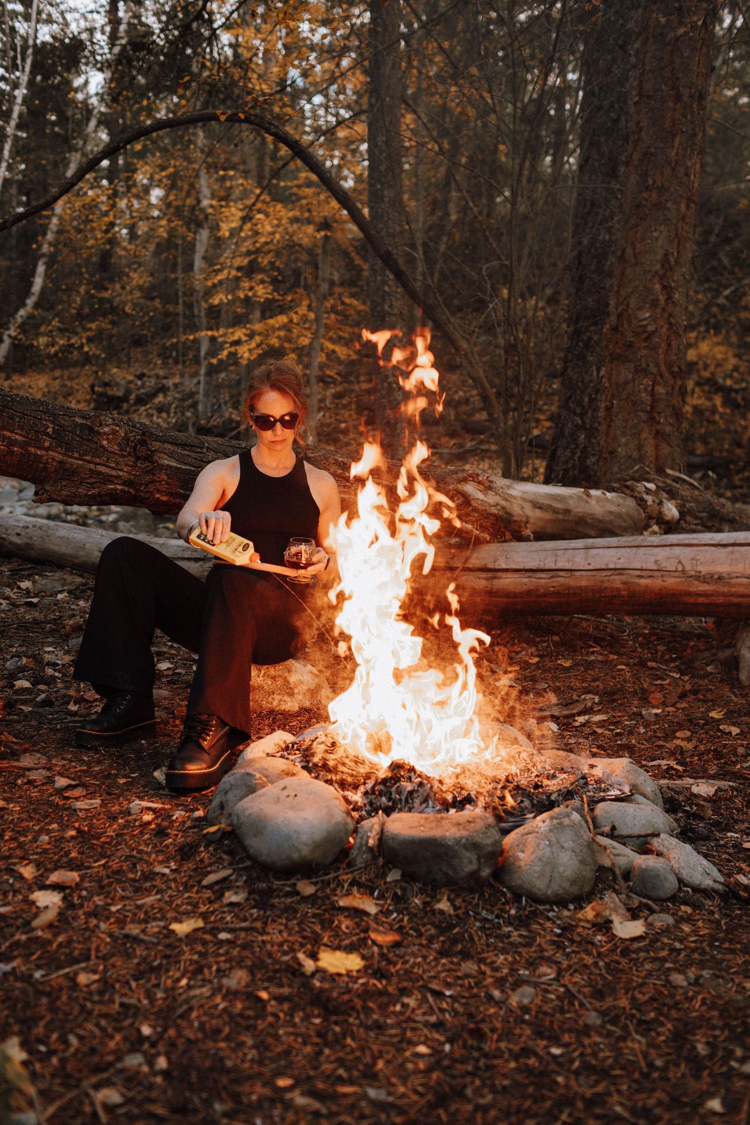 A cozy autumn campfire scene with someone sitting on a log beside a stone-ringed fire in the woods at dusk.
