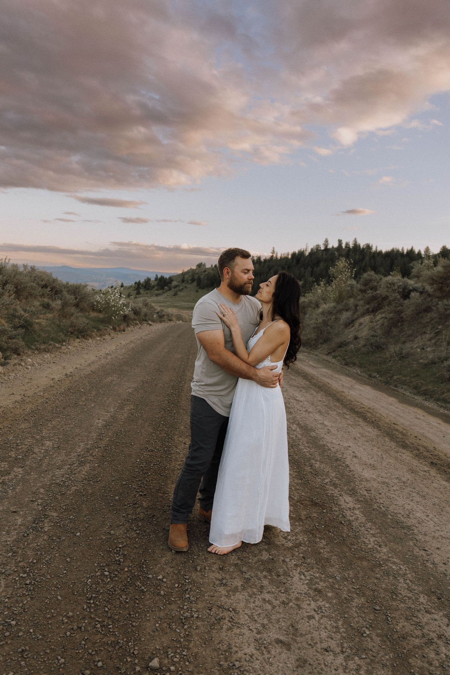 A couple embraces on a dirt road at sunset with mountains and dramatic clouds in the background.