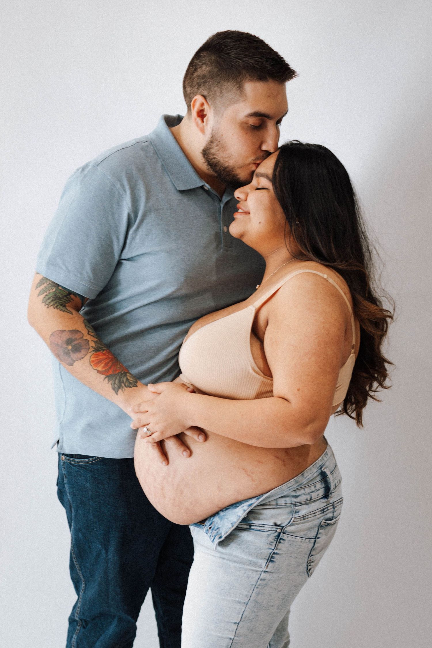 An intimate maternity photo showing couple embracing, with man in blue polo and woman in crop top and jeans.