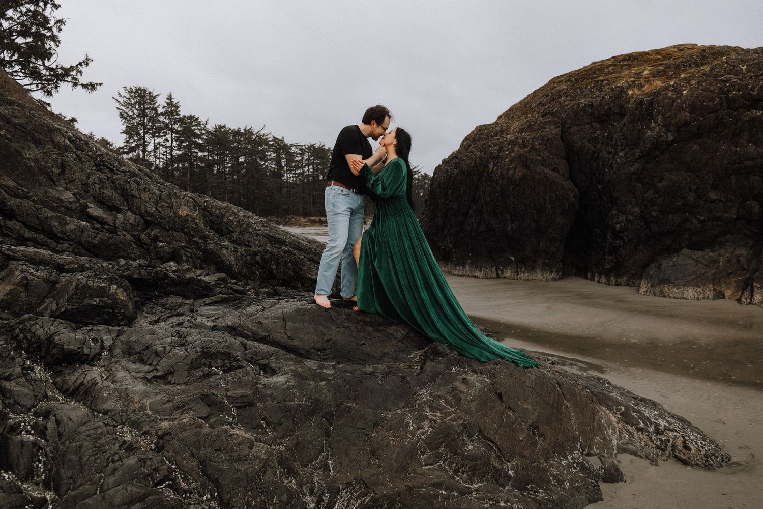 Couple kissing on rocky beach, woman in flowing emerald green gown, moody coastal scenery with trees and cliffs.
