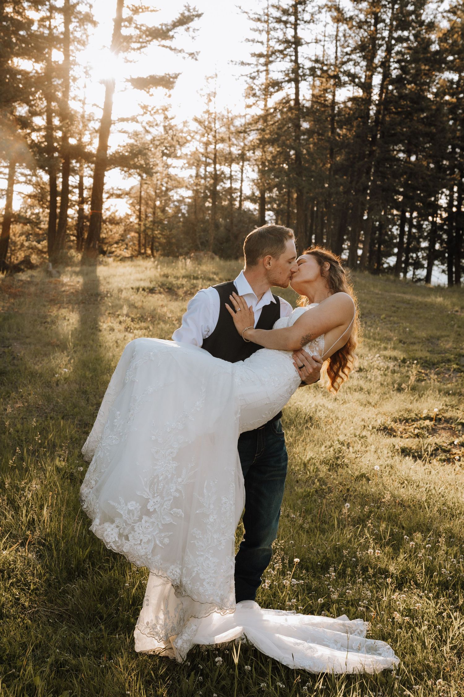 Groom lifts bride in white lace gown for a kiss in a sunlit forest clearing during golden hour wedding photos.