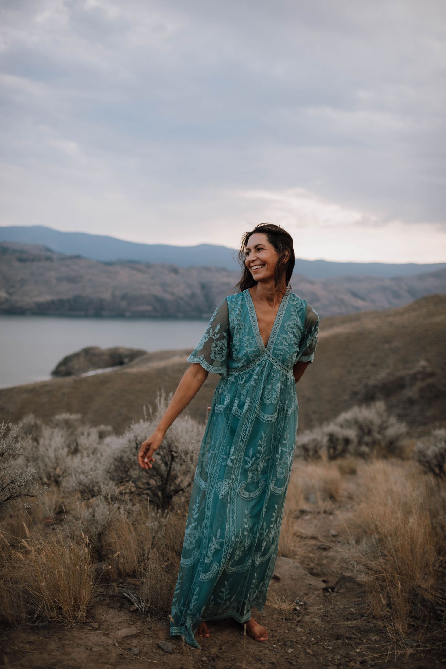 A person in a flowing turquoise dress stands on a hillside overlooking a serene lake.