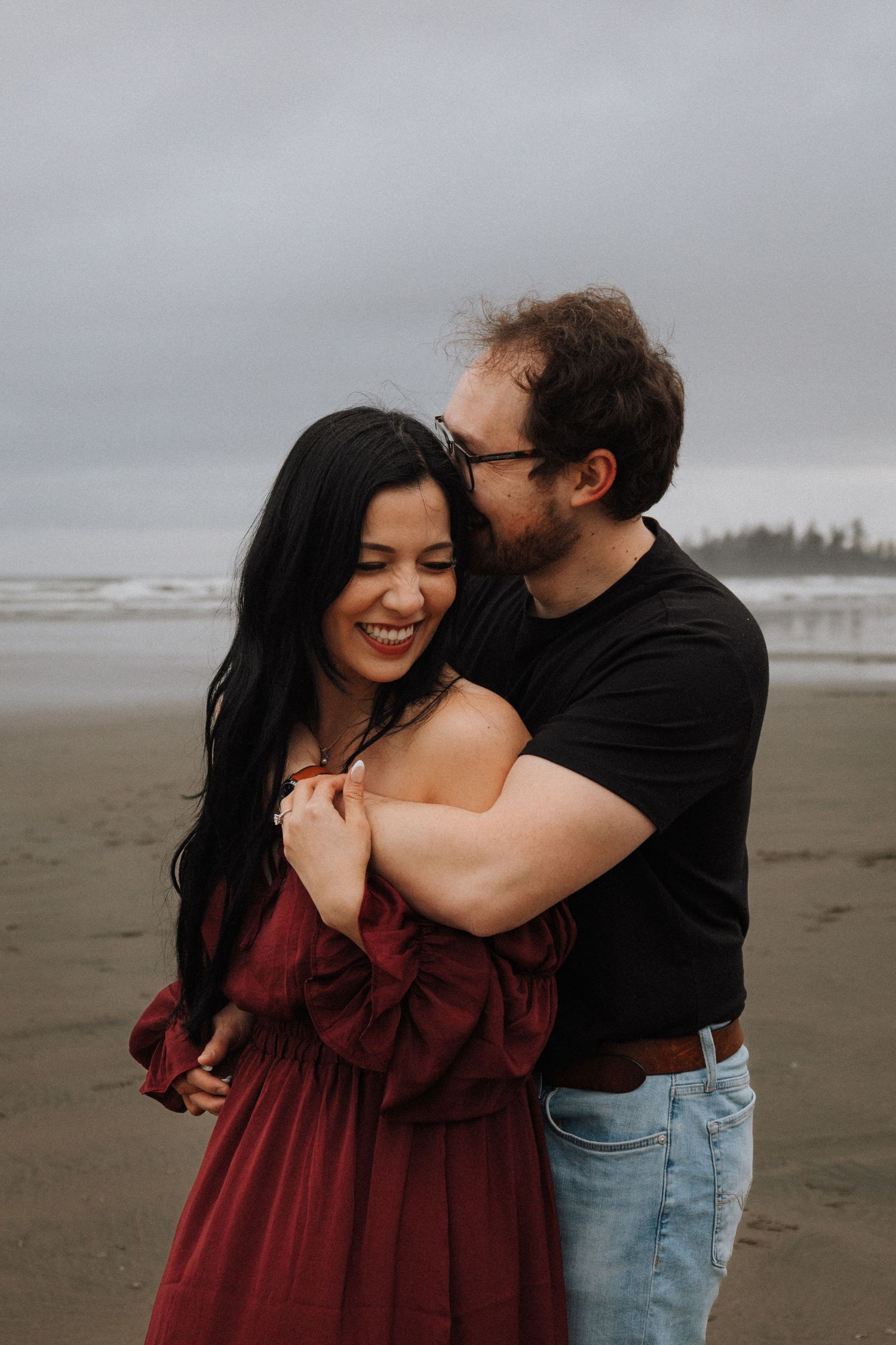 Couple embracing on a cloudy beach, woman in red dress laughing as man kisses her cheek. Long Beach BC Tofino