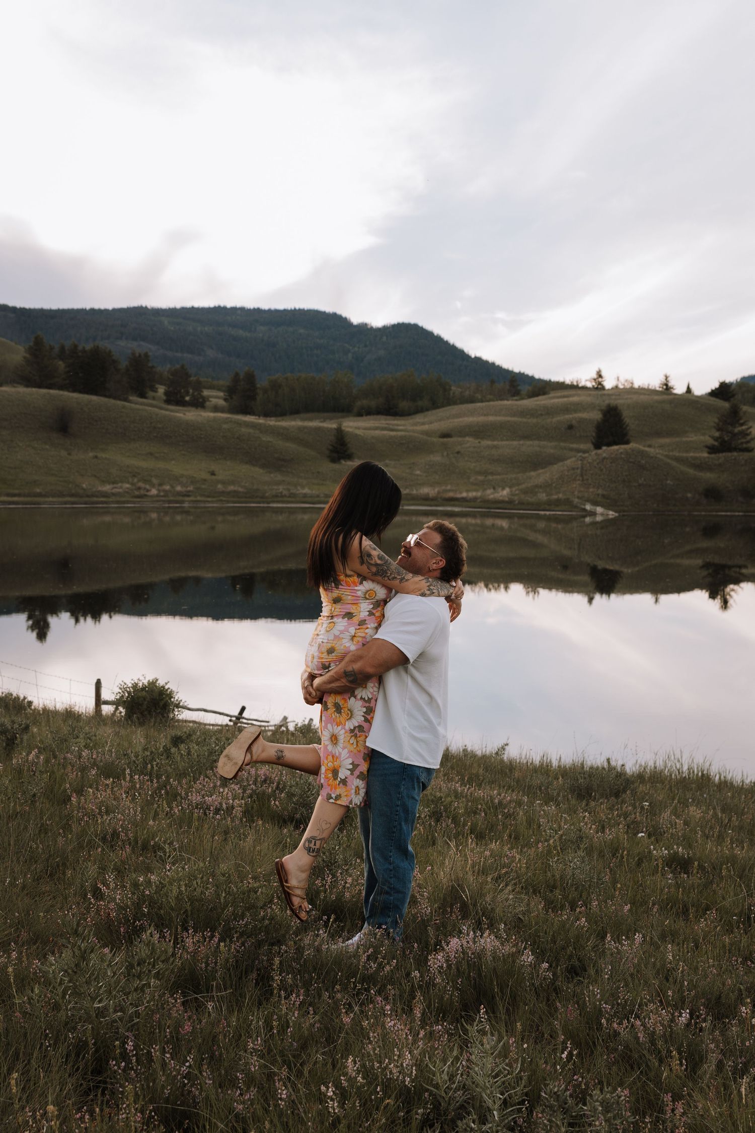 Couple embracing by a serene mountain lake, man lifting woman in floral dress, lush green hills reflected in calm water.