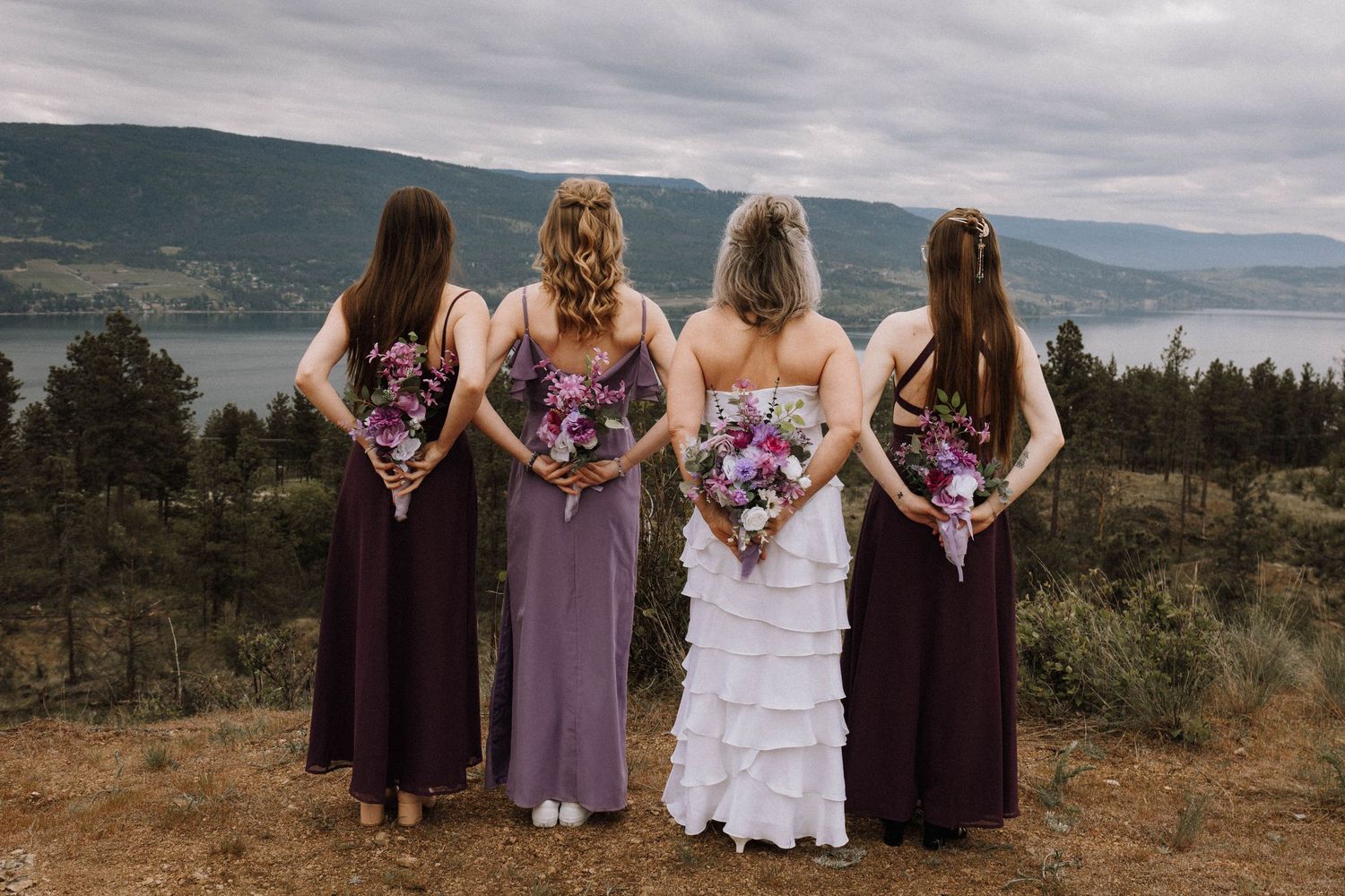 A bridal party in purple dresses and white gown stands overlooking a scenic mountain lake vista.