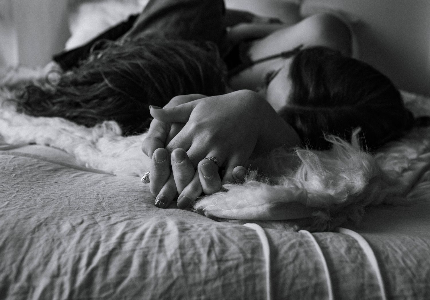 Black and white image of intertwined hands resting on a bed.