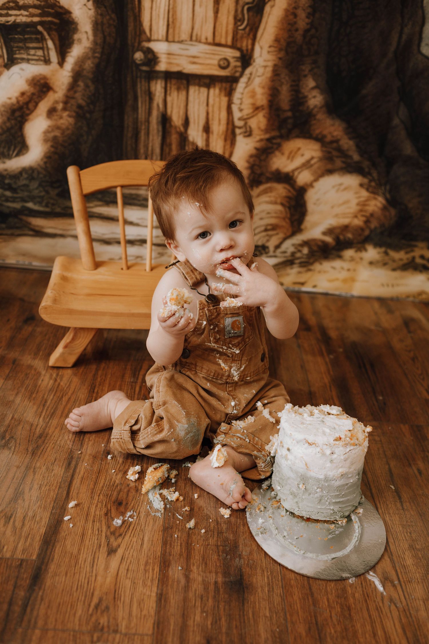 Baby in overalls enjoying smash cake on wooden floor with vintage backdrop.