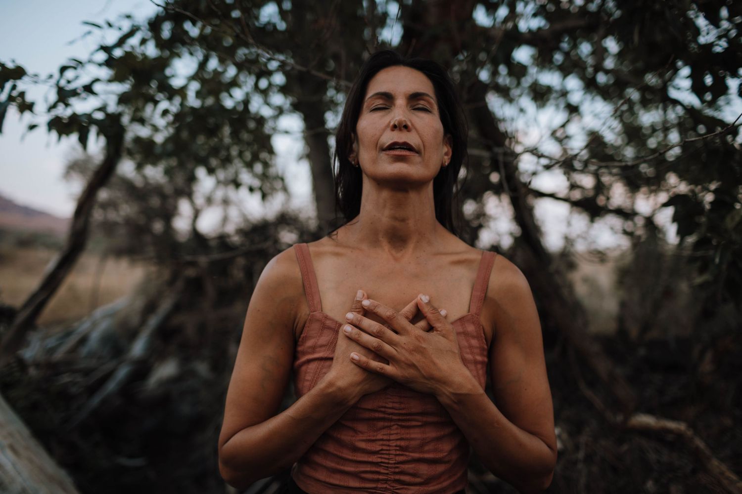 An atmospheric outdoor portrait with hands held to heart against a natural background.