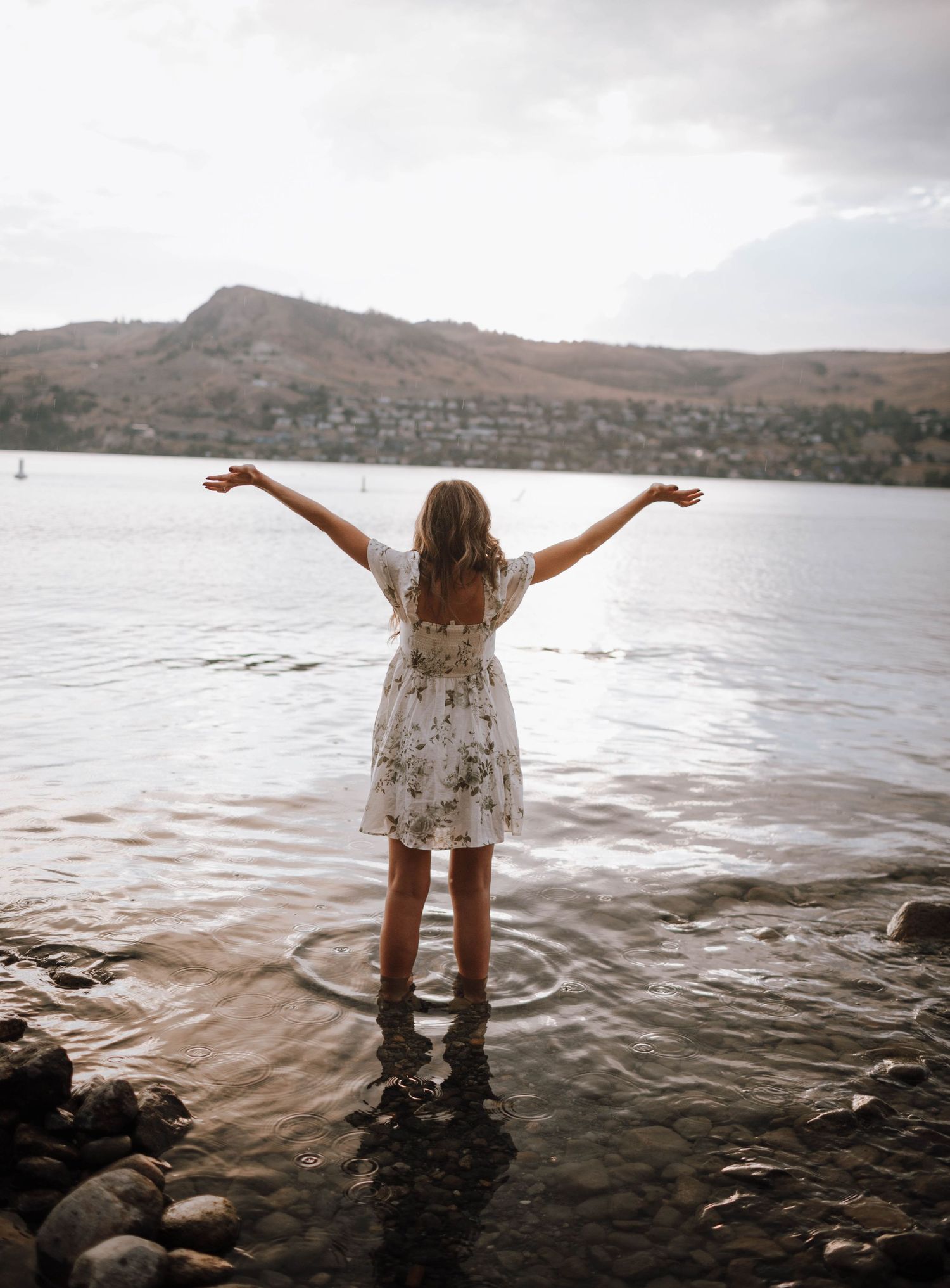 Person in white dress stands in lake water with arms outstretched against mountainous backdrop.