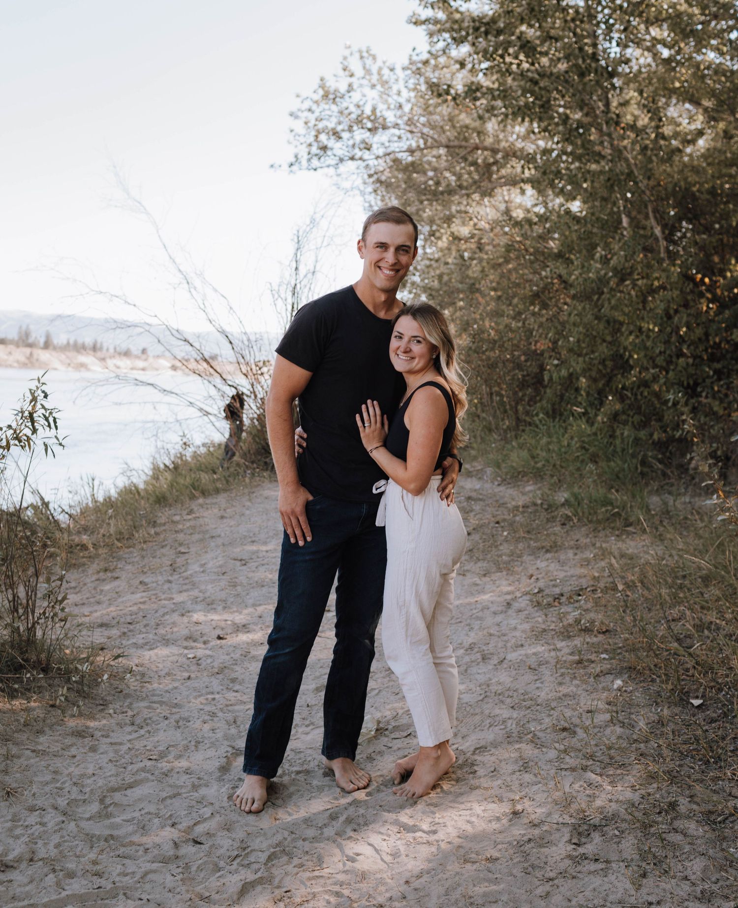 A couple poses together on a scenic dirt trail overlooking water and mountains.