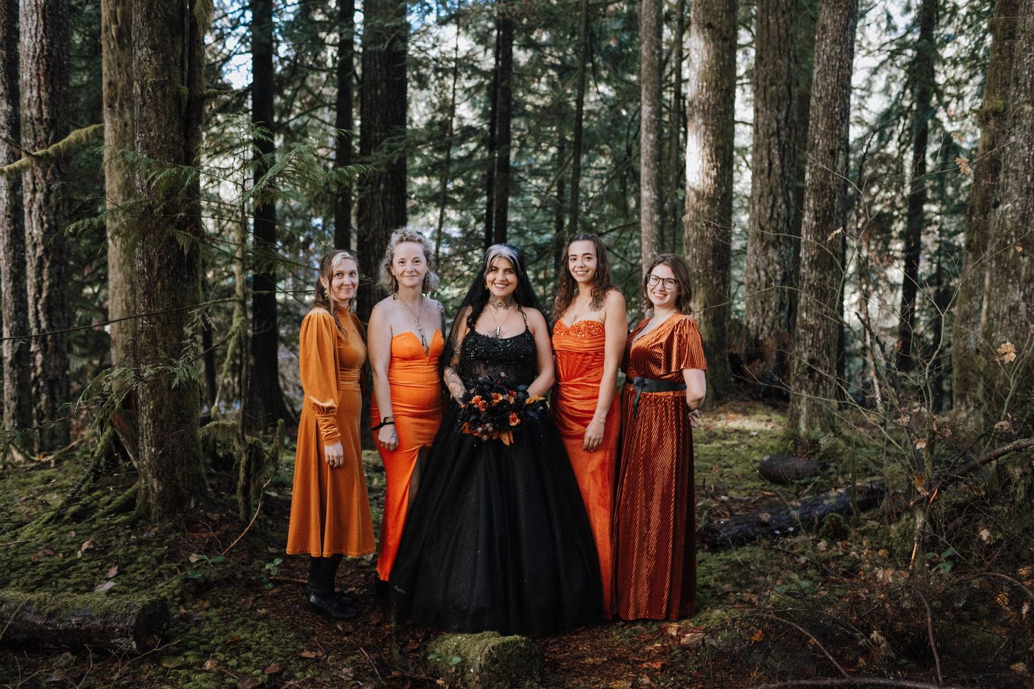 Wedding party in contrasting orange dresses and black gown pose together in a moody forest setting.