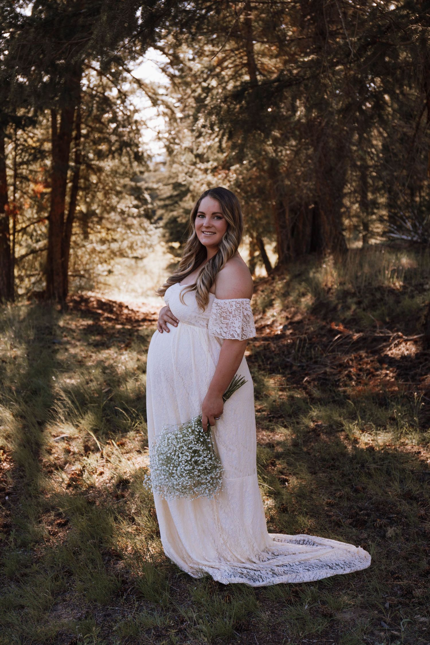 Ethereal maternity photograph in a sunlit forest featuring an off-shoulder white lace gown and delicate baby's breath bouquet.