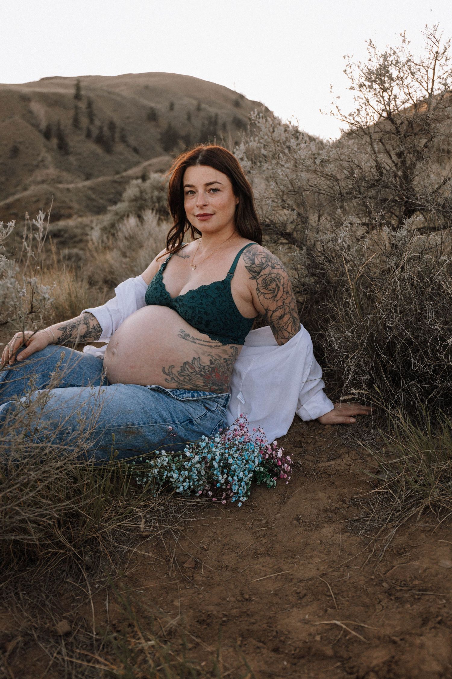 A person reclines on brown desert hills at sunset wearing a white top and jeans amidst wild brush and shrubs.