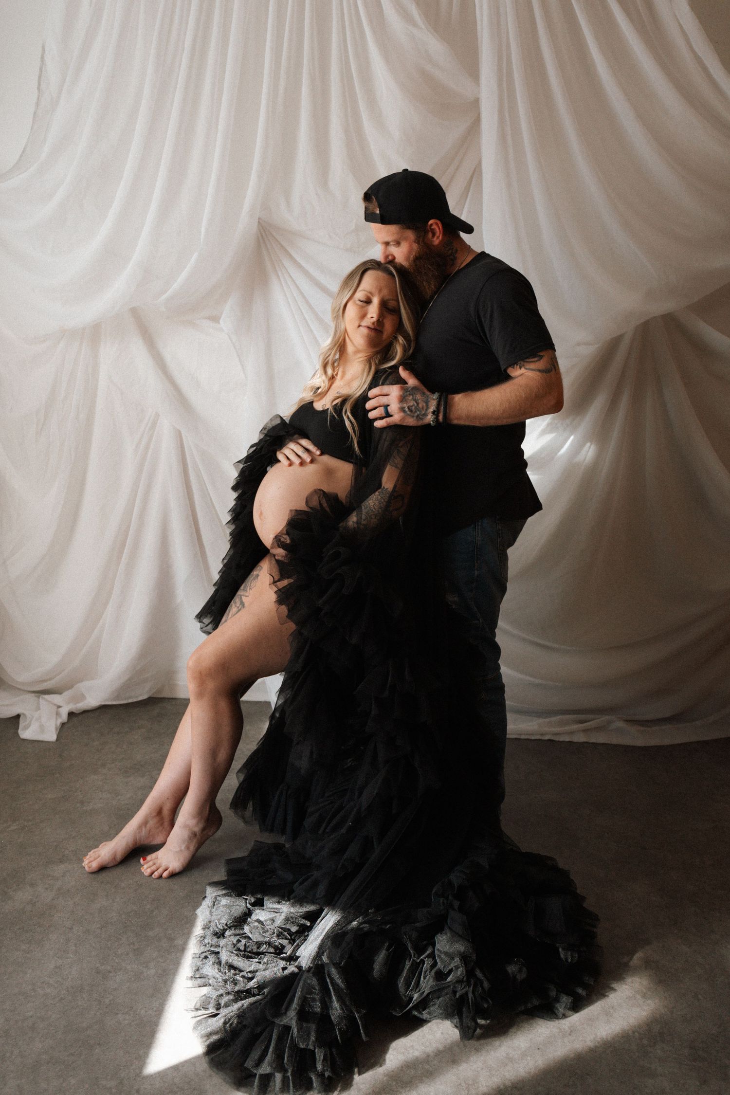 A couple in black attire poses intimately against a white draped backdrop in a romantic photoshoot series.