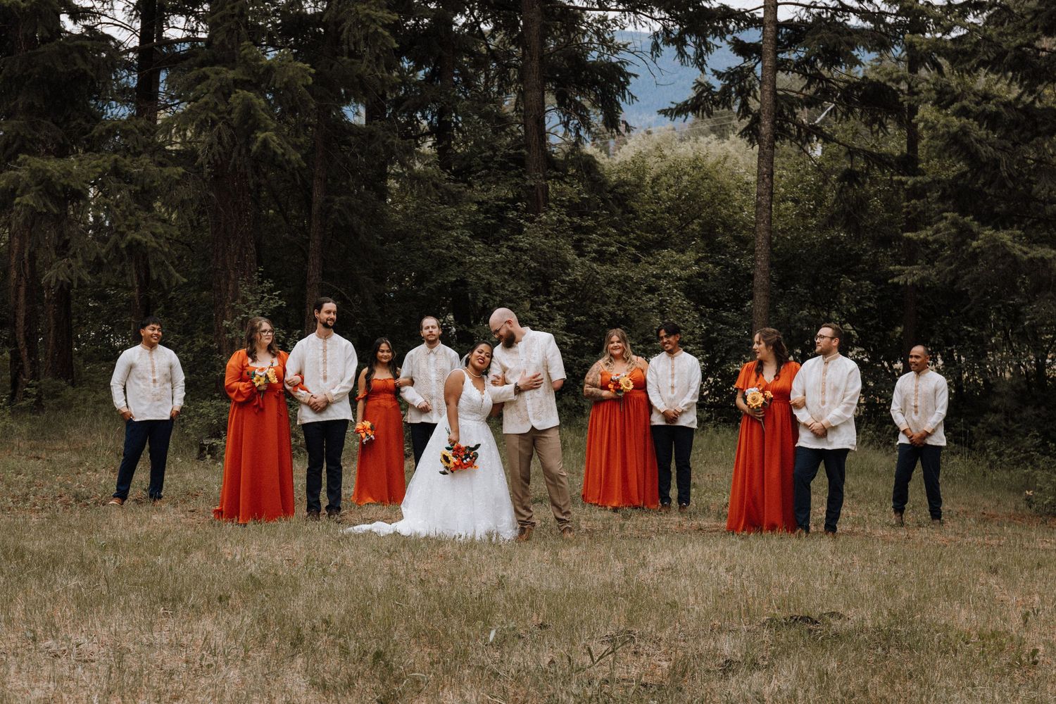 Wedding party in white and red attire poses in a rustic outdoor field setting with tall grass and trees.