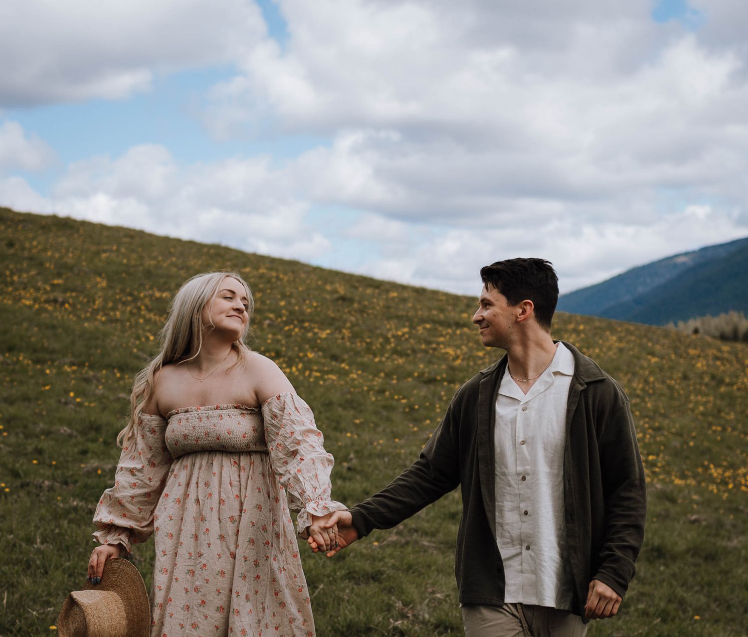 A couple holds hands while walking through a scenic mountain meadow filled with wildflowers on a cloudy day.