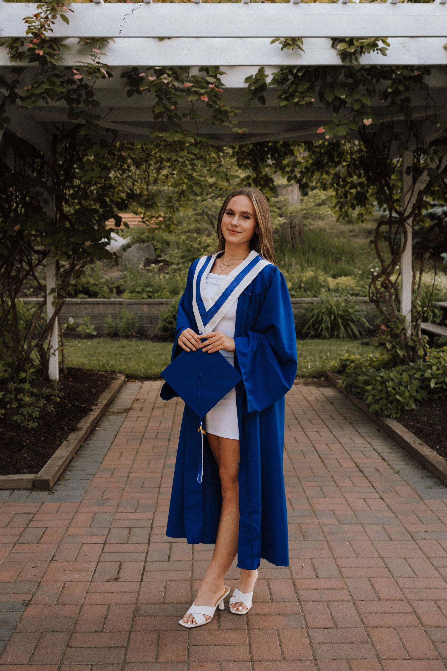 Young woman in blue graduation gown and cap posing on brick path under pergola surrounded by lush greenery.