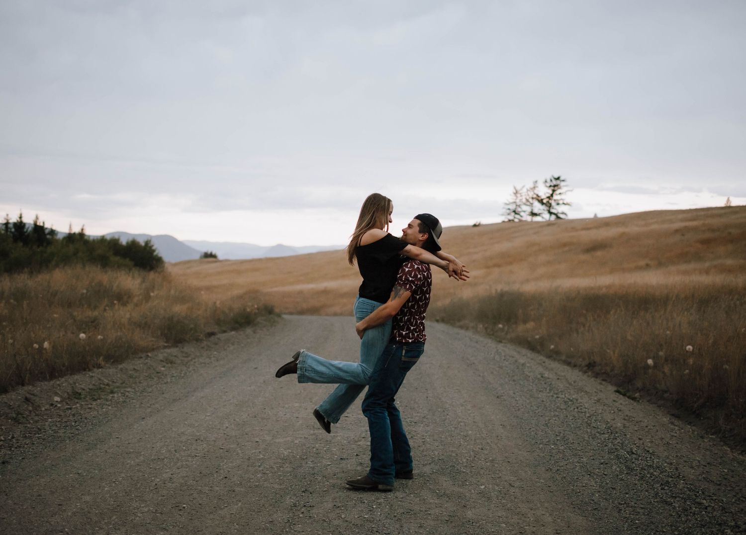 A playful couple spins and dances on a remote dirt road surrounded by rolling hills.