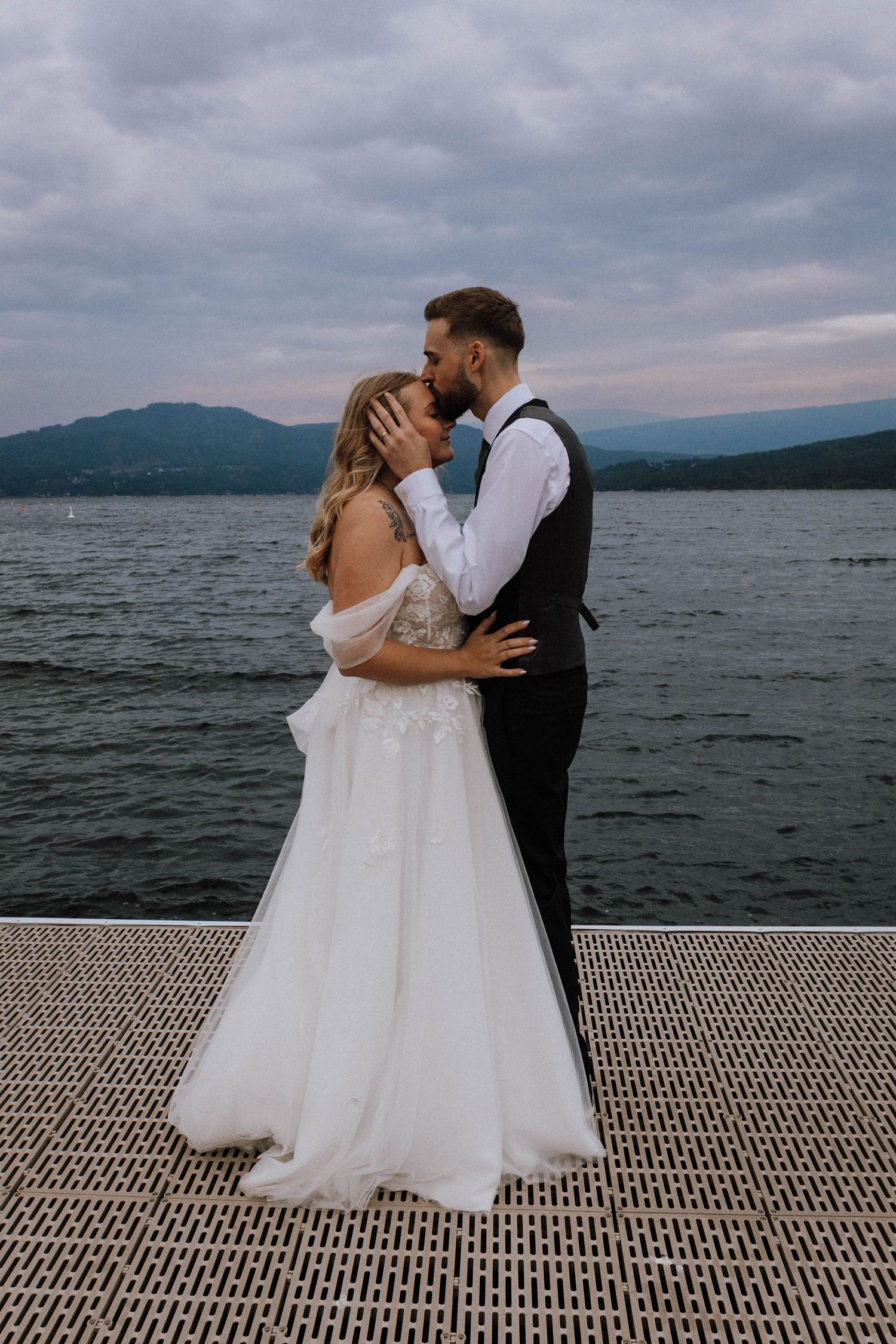 A romantic wedding couple shares a kiss on a dock overlooking a misty mountain lake.