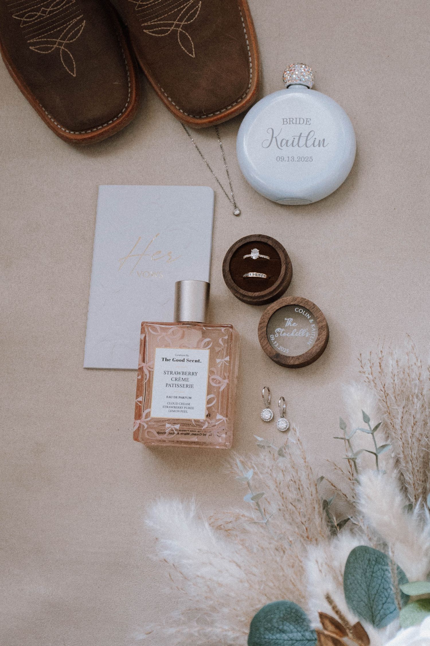 Wedding details flatlay with perfume bottles, jewelry, and dried pampas grass on a neutral beige background.