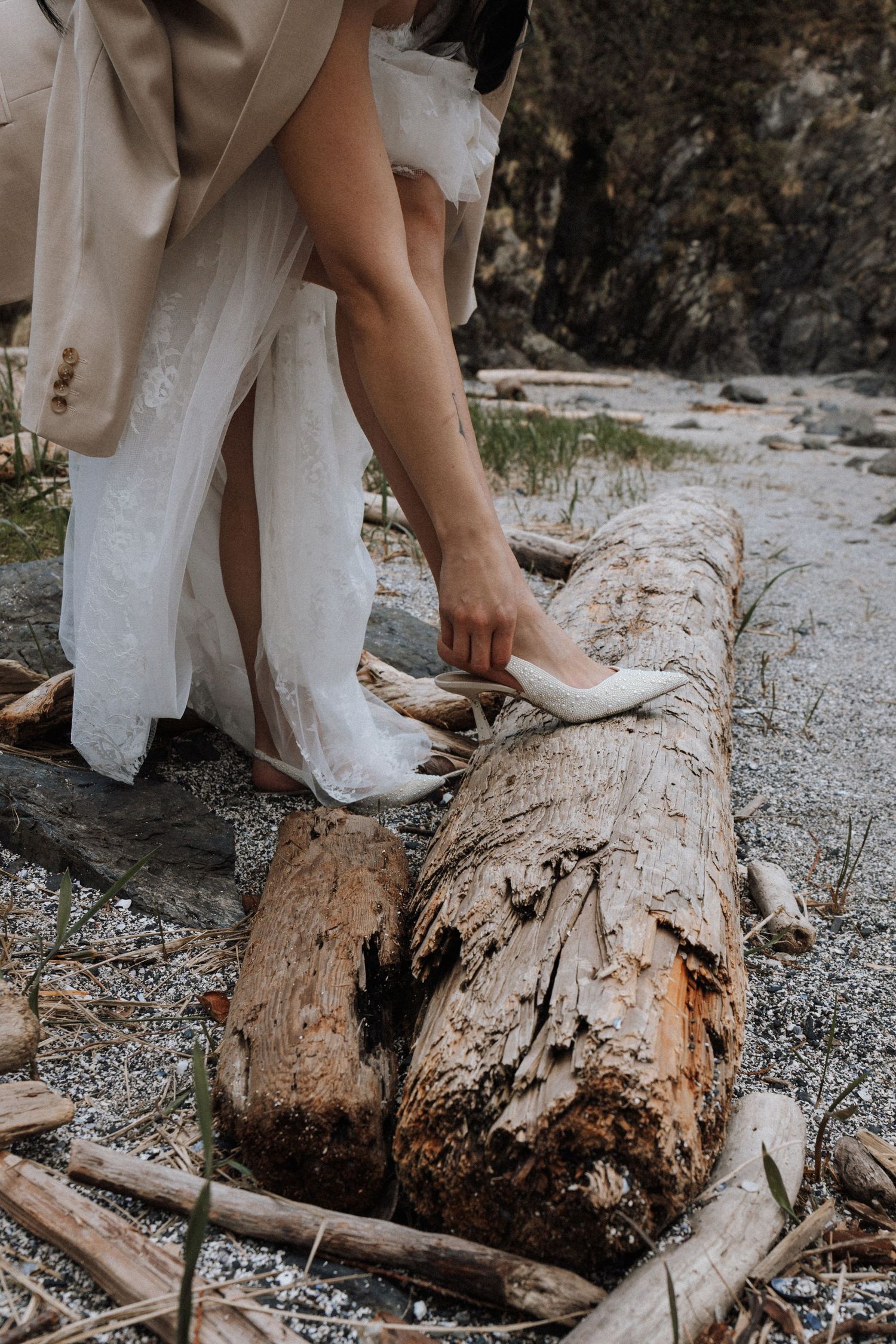 Bride in lace gown placing white heels on a driftwood log on a rocky beach.