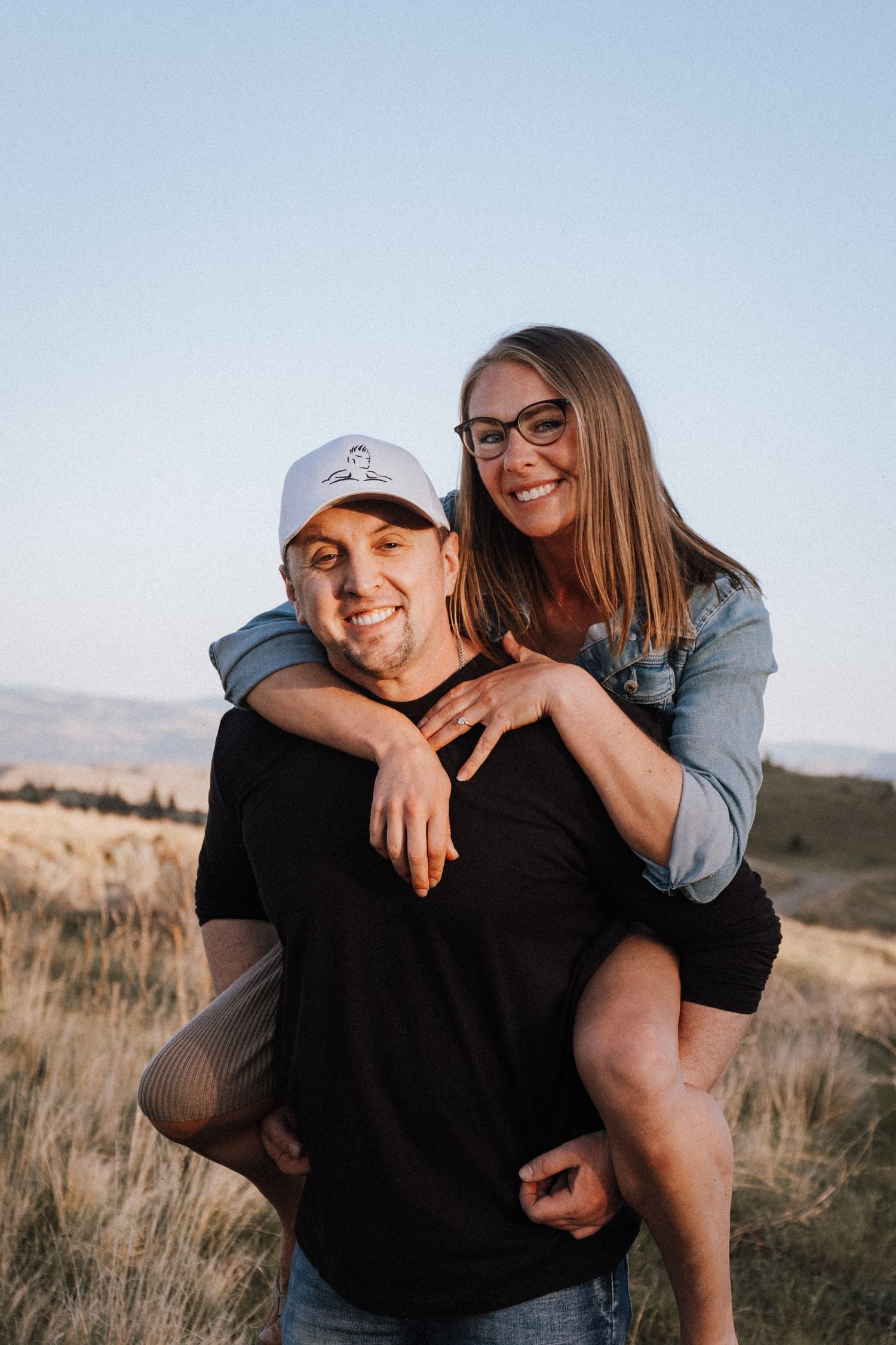 A smiling couple outdoors, man in white cap and black tee giving woman in glasses a piggyback ride in a golden field.