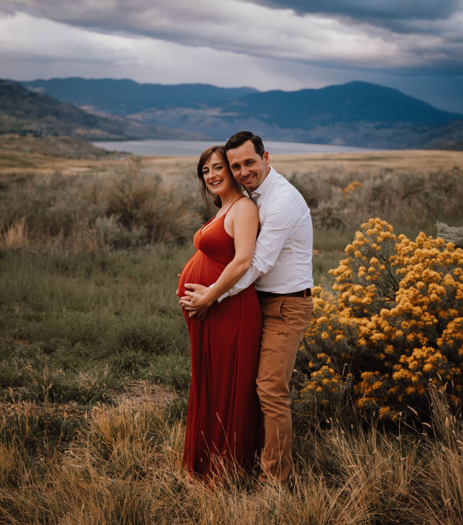 Couple embraces in front of mountain vista with dramatic clouds and golden rabbitbrush flowers.