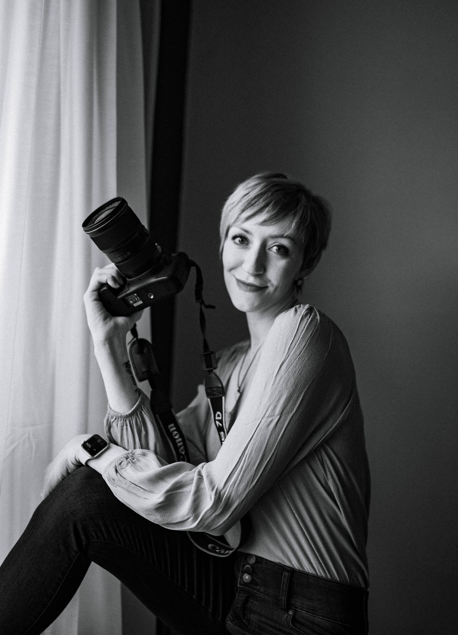 A black and white portrait of a photographer holding a professional camera by a window.