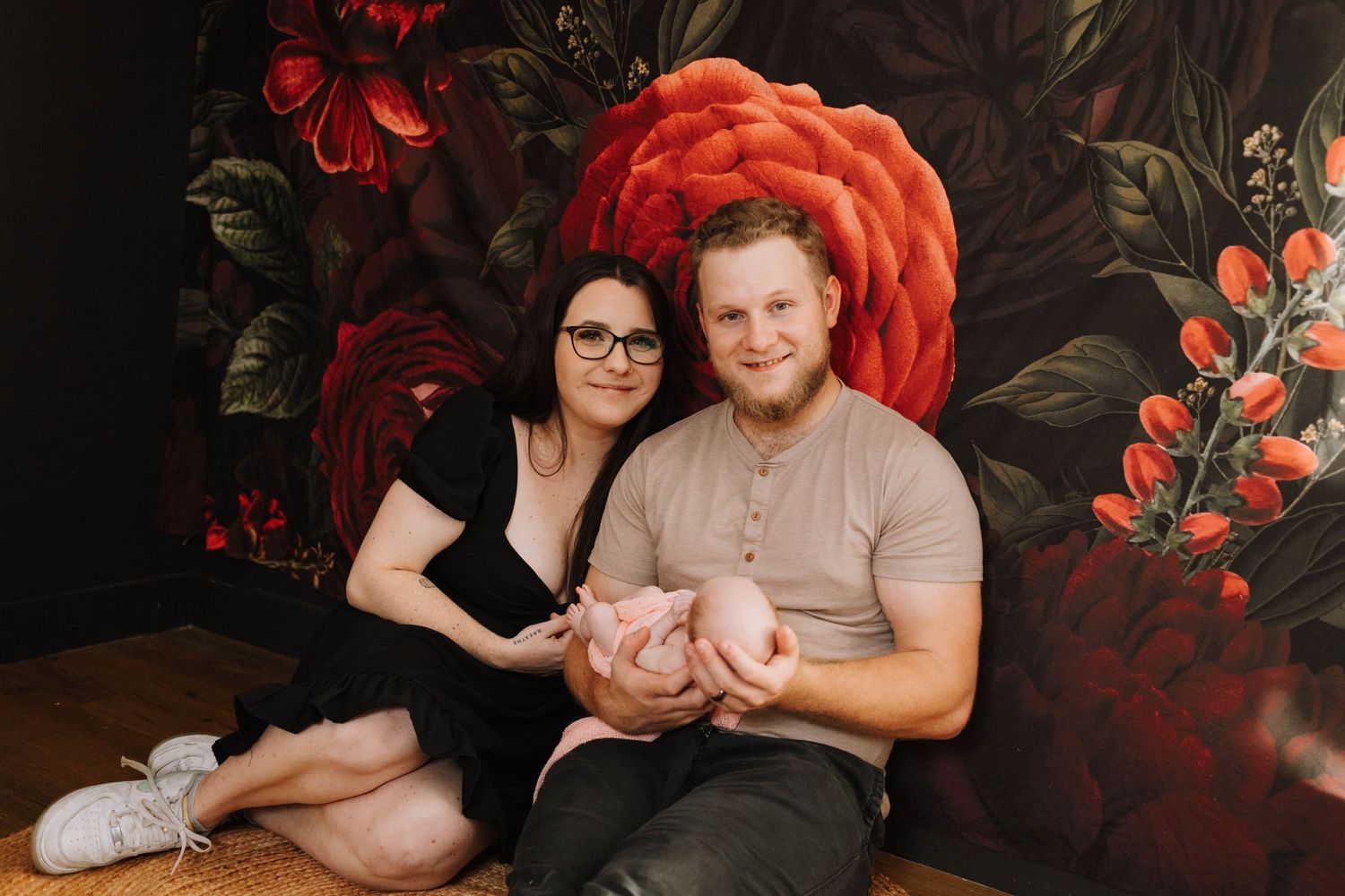 Family portrait against dramatic floral wallpaper holding newborn baby.