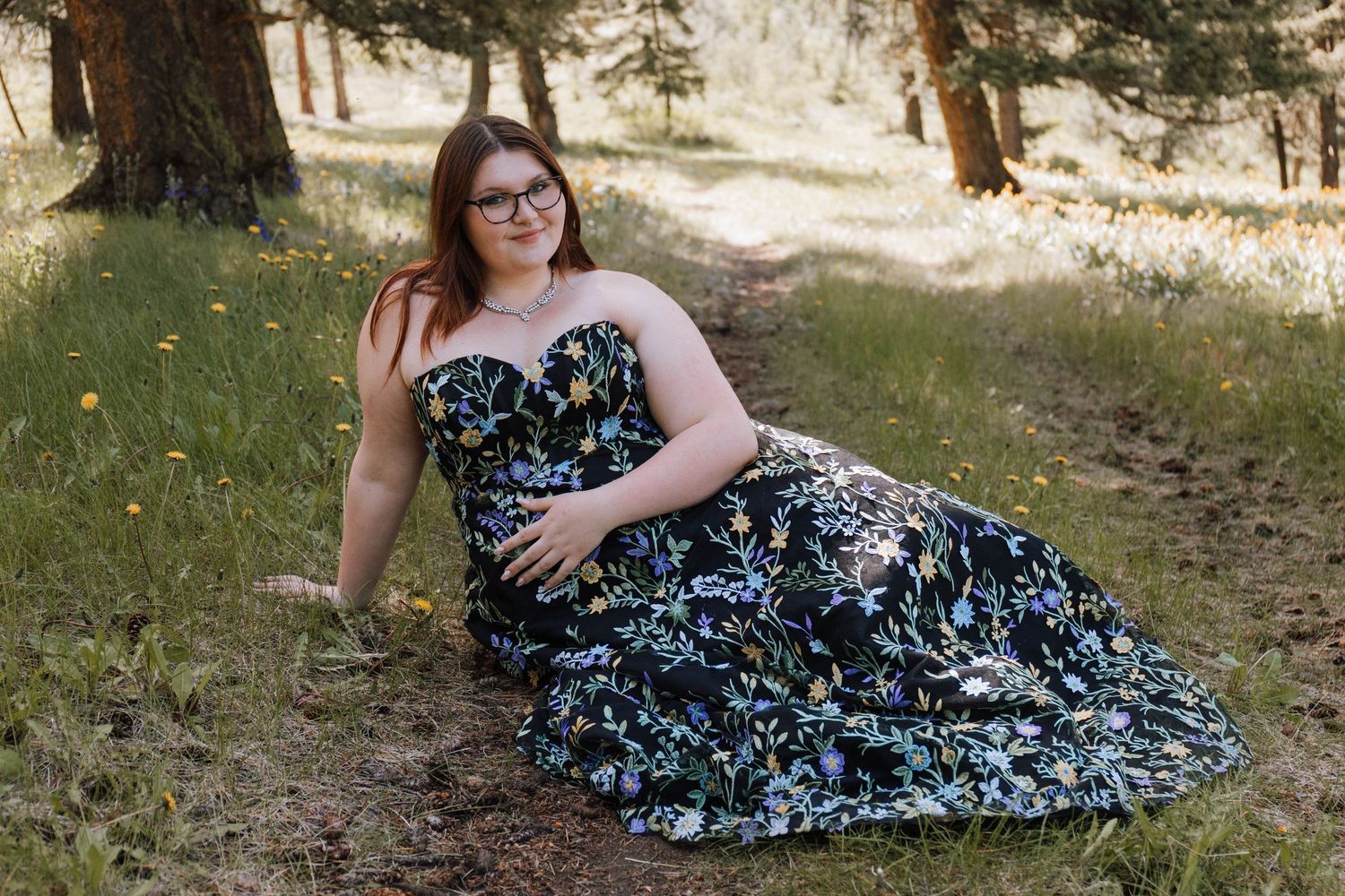 Someone in glasses reclines on grassy ground wearing a strapless floral dress in a woodland setting.