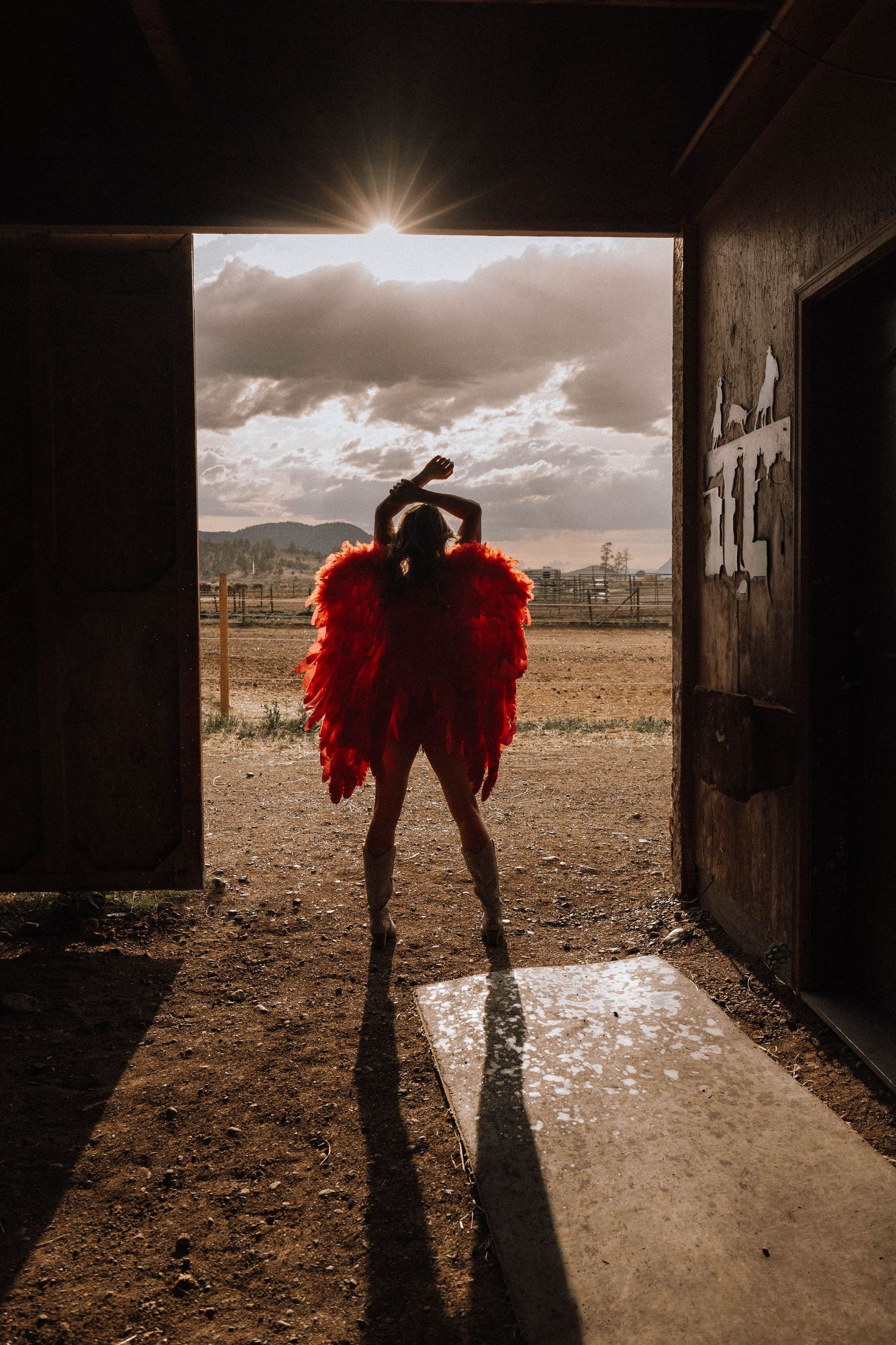 Silhouetted figure in dramatic red wings stands in barn doorway as sunlight streams in over rustic landscape.