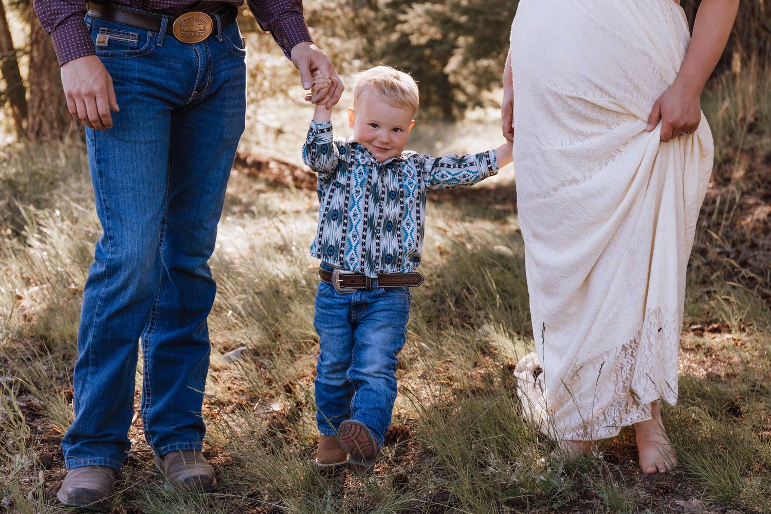Toddler in blue jeans and patterned shirt holds hands with parents while standing in grassy outdoor setting at sunset.