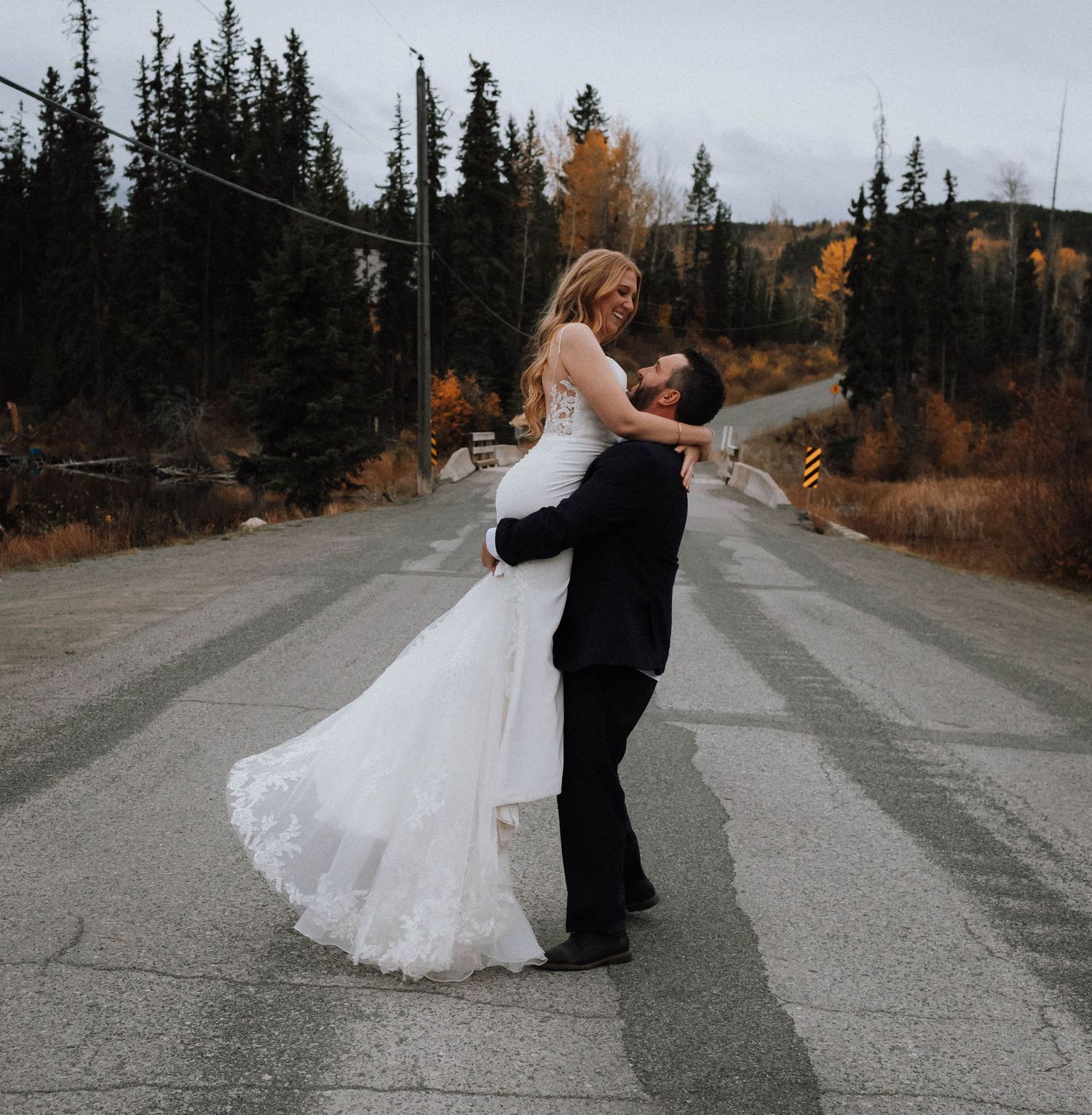 A couple in wedding attire dances and twirls on a mountain road surrounded by autumn trees and evergreens.
