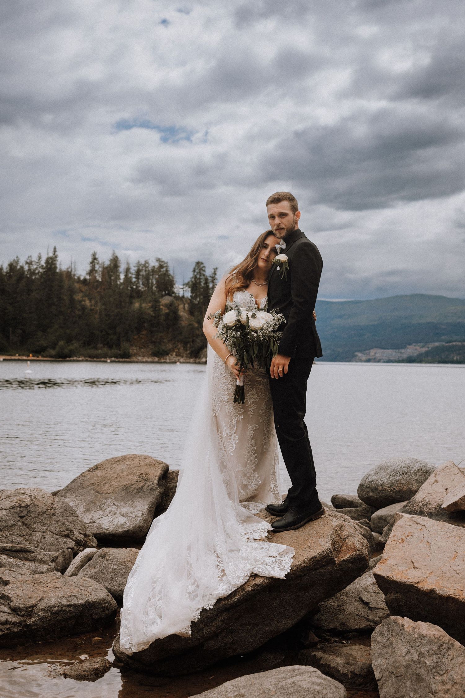 Bride and groom standing on rocky shoreline with lake and mountains backdrop on cloudy wedding day.